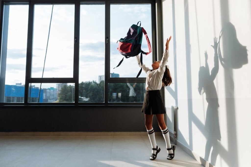 A joyful girl in school uniform tossing her backpack in a sunlit classroom.